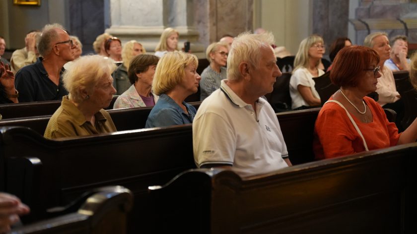 Na zdjęciu widzimy grupę osób siedzących w ławkach kościelnych, uczestniczących w uroczystości. Wśród widocznych postaci znajdują się głównie starsze osoby, które są skupione i uważnie słuchają lub obserwują przebieg wydarzenia. Na pierwszym planie widoczna jest starsza kobieta w beżowej kurtce oraz mężczyzna w białej koszulce polo. W tle, w głębi kadru, znajdują się inni uczestnicy, którzy również są skupieni na ceremonii.