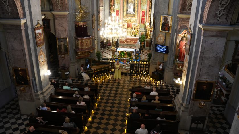 Zdjęcie przedstawia wnętrze kościoła widziane z perspektywy z góry, prawdopodobnie z balkonu lub chóru. Na pierwszym planie widać wiernych siedzących w ławkach, a na drugim planie scenę przed ołtarzem, na której odbywa się występ muzyczny. Na środku stoi kobieta w żółtej sukni, trzymająca nuty, prawdopodobnie śpiewająca, obok niej widoczny jest klawesyn, przy którym siedzi inna osoba grająca na instrumencie. W tle widać bogato zdobiony ołtarz, oświetlony licznymi świecami, co dodaje uroczystego klimatu całemu wydarzeniu. Świece również ustawione są na podłodze wzdłuż przejścia, co tworzy niezwykły nastrój.