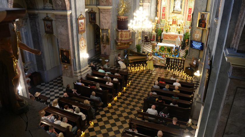 Zdjęcie zostało wykonane z wysokości, najprawdopodobniej z balkonu lub podestu wewnątrz kościoła. Widzimy z góry nawy głównej z ustawionymi ławkami, w których siedzą uczestnicy wydarzenia. Na środku nawy, przed ołtarzem, znajduje się artystka ubrana w żółtą suknię, prawdopodobnie śpiewająca. Obok niej, po lewej stronie, osoba gra na klawesynie, który jest pięknie zdobiony. Przed artystką na posadzce ustawiono liczne zapalone świece, które tworzą przyjemny, kameralny nastrój. W tle widać bogato zdobiony ołtarz oraz dekoracje z kwiatami. Zdjęcie pokazuje uroczystość lub koncert, który odbywa się w kościele, a jego uczestnicy skupieni są na występie. Ujęcie z góry pozwala zobaczyć cały kontekst wydarzenia oraz jego oprawę artystyczną.