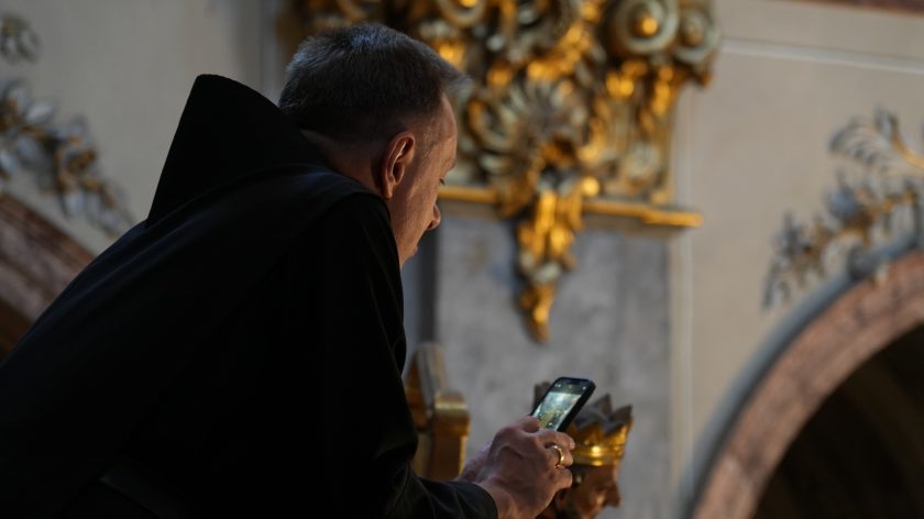 Widok z boku na zakonnika w czarnej szacie, który trzyma w rękach smartfon i robi zdjęcie wnętrza kościoła. W tle widać złote detale ołtarza