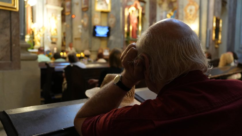 Starszy mężczyzna, widziany od tyłu, opiera głowę na dłoni, wpatrując się w ołtarz podczas wydarzenia w kościele. Światło świec delikatnie rozświetla wnętrze.