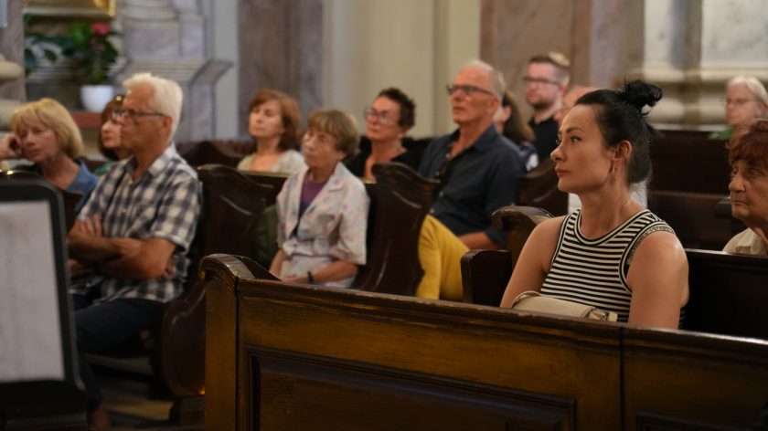 Grupa ludzi siedzi w ławkach w kościele, wszyscy uważnie słuchają. Na pierwszym planie kobieta w biało-czarnym paskowanym stroju.