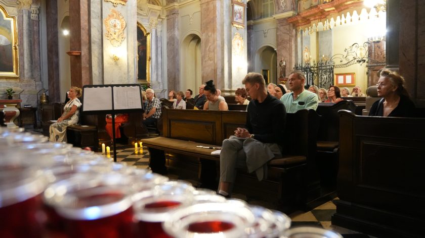 Szerszy kadr na wnętrze kościoła z widokiem na kilka rzędów świec, które są na pierwszym planie. W tle siedzą wierni, słuchający występu.