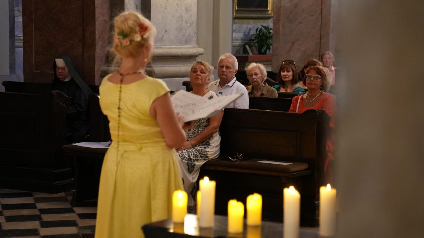 Kobieta w żółtej sukni stoi tyłem do aparatu, trzymając otwarty śpiewnik. Przed nią siedzący ludzie w ławkach, patrzący w jej kierunku. Na dalszym planie widać świece i zakonnicę w habicie.