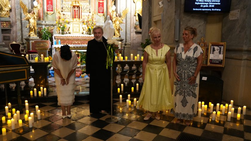 Grupa czterech osób stoi przed ołtarzem, dwie kobiety w eleganckich sukniach, mężczyzna w sutannie oraz kobieta w białym stroju kłaniająca się.