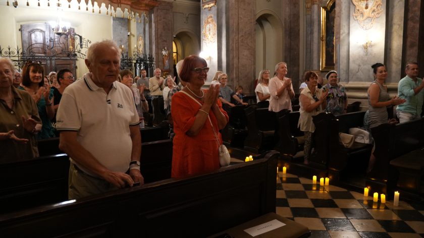 Publiczność stojąca w ławkach kościelnych, oklaskująca na zakończenie wydarzenia. Na pierwszym planie starszy mężczyzna w białej koszulce i kobieta w czerwonej sukience, a w tle widać innych uczestników wydarzenia, którzy również biją brawo.