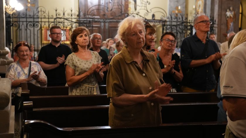 Zbliżenie na grupę widzów klaskających podczas zakończenia wydarzenia. Starsza kobieta w zielonej sukience i mężczyzna w okularach znajdują się na pierwszym planie, z wyrazami uznania na twarzach.