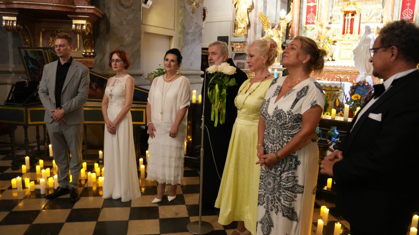 Na zdjęciu widzimy grupę sześciu osób, ustawionych w rzędzie, podczas uroczystości w kościele. Od lewej do prawej stoją: młody mężczyzna w szarym garniturze, młoda kobieta w długiej białej sukni, kobieta w eleganckiej białej sukience z długim naszyjnikiem z pereł, mężczyzna w czarnym stroju duchownym trzymający bukiet białych róż, kobieta w żółtej sukni oraz kobieta w białej sukni z czarnymi wzorami. W tle widać bogato zdobione wnętrze kościoła z licznymi świecami ustawionymi na podłodze oraz ołtarzem udekorowanym kwiatami i świętymi figurami.