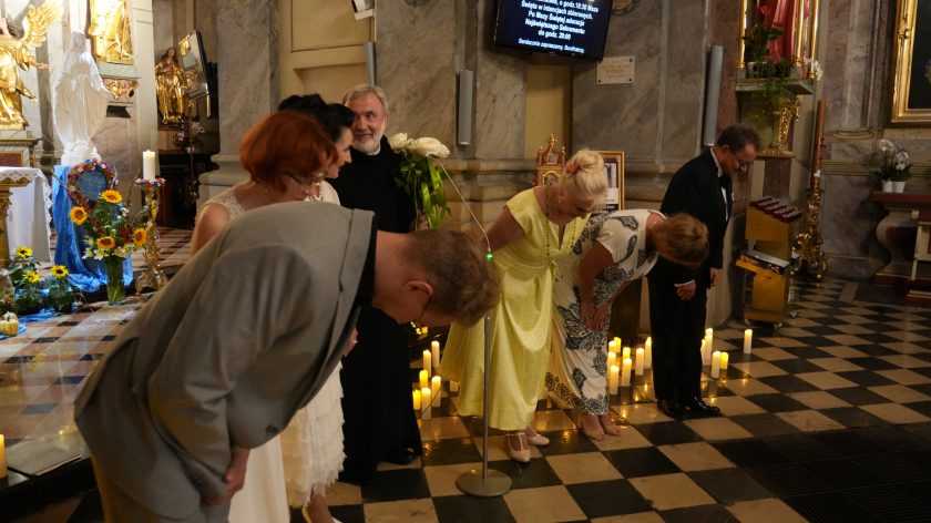 Na zdjęciu widzimy grupę ludzi, którzy składają ukłon, co sugeruje zakończenie uroczystości lub koncertu, podczas którego otrzymali wyrazy uznania od publiczności. Na pierwszym planie widać kobietę w jasnej sukience i mężczyznę w garniturze, a w tle kolejne osoby, również uczestniczące w ukłonie. Scena ma miejsce w kościele, co można wywnioskować z widocznych elementów sakralnych, takich jak figury i kwiatowe dekoracje. Świeczki ustawione na podłodze dodają atmosfery powagi i skupienia. Cała scena emanuje uroczystym charakterem.