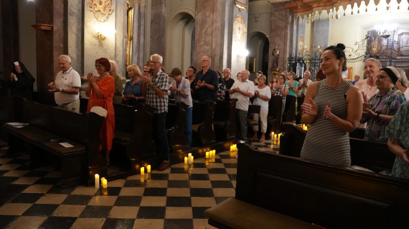 Na zdjęciu widzimy grupę osób stojących w kościele, które wyglądają na zaangażowane w uroczystość lub wydarzenie. Wszyscy są zwróceni w jednym kierunku i klaszczą, co sugeruje, że uczestniczą w zakończeniu występu lub ceremonii. W pierwszym rzędzie po lewej stronie widzimy starszego mężczyznę w białej koszulce oraz kobietę w pomarańczowej sukience. Wśród uczestników jest także siostra zakonna stojąca po lewej stronie w czarno-białym habicie. Cała scena rozgrywa się w kościele o bogato zdobionym wnętrzu z marmurowymi kolumnami i żółtymi świecami ustawionymi na podłodze, co nadaje całości uroczysty i podniosły nastrój.