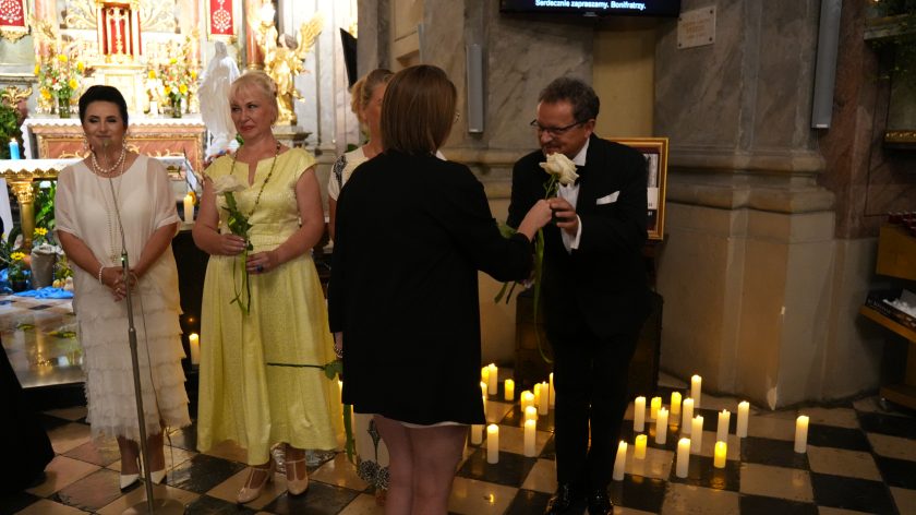 Fotografia przedstawia grupę elegancko ubranych osób - kobiet i mężczyzn, pozujących do zdjęcia w kościele na tle ołtarza. Wszystkie osoby trzymają w rękach po jednej, białej róży. Wśród osób znajduje się młoda kobieta wręczająca kwiat jednemu z mężczyzn. Przed ołtarzem widać rozstawione, zapalone świece.