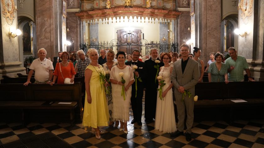 Fotografia przedstawia grupę elegancko ubranych osób - kobiet i mężczyzn, pozujących do zdjęcia w kościele na tle zabytkowego wejścia. Wszystkie osoby trzymają w rękach po jednej, białej róży. Wśród osób znajduje się brat zakonny. W tle widać inne osoby stojące w ławach kościelnych.