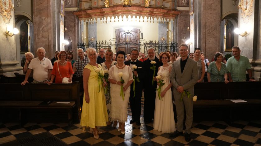 Fotografia przedstawia grupę elegancko ubranych osób - kobiet i mężczyzn, pozujących do zdjęcia w kościele na tle zabytkowego wejścia. Wszystkie osoby trzymają w rękach po jednej, białej róży. Wśród osób znajduje się brat zakonny. W tle widać inne osoby stojące w ławach kościelnych.