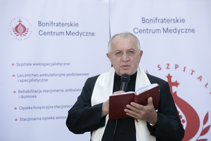 Duchowny w czarnej sutannie trzyma mikrofon i otwartą księgę, przemawiając do zgromadzonych. W tle znajduje się baner "Bonifraterskie Centrum Medyczne" z informacjami o działalności placówki