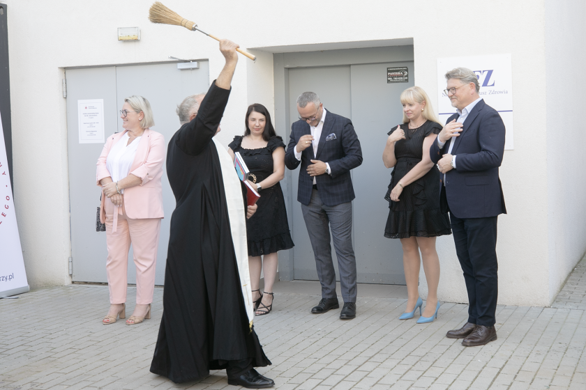 Duchowny w czarnej sutannie błogosławi grupę osób na zewnątrz budynku, używając miotły. Ludzie stoją w półokręgu, niektórzy wykonują znak krzyża. W tle widoczne są drzwi i tablica informacyjna Narodowego Funduszu Zdrowia