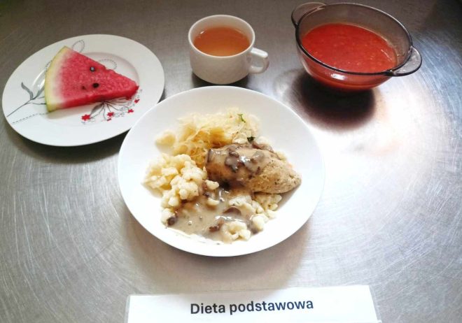 Zdjęcie z obiadem oznaczone "dieta podstawowa": Na zdjęciu widoczny jest posiłek obiadowy ułożony na metalowej powierzchni. W centrum znajduje się biały talerz z głównym daniem, na którym podano kluseczki z sosem, kawałek mięsa w sosie pieczeniowym oraz porcję kiszonej kapusty. Po lewej stronie talerza znajduje się deser w postaci trójkąta arbuza na białym talerzyku. Po prawej stronie znajduje się miseczka z czerwoną zupą, a powyżej filiżanka z przezroczystym napojem (prawdopodobnie kompotem). Na dole zdjęcia widoczna jest karteczka z napisem "Dieta podstawowa".