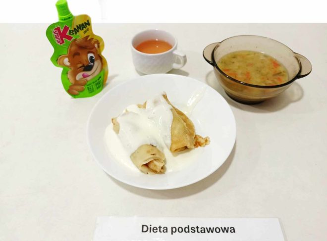 Taca z posiłkiem oznaczona jako "Dieta podstawowa". Na białym talerzu znajdują się naleśniki polane białym sosem, prawdopodobnie śmietaną. Obok znajduje się miska z zupą warzywną, filiżanka z różowym napojem oraz opakowanie w kształcie misia z musem owocowym przeznaczonym dla dzieci.