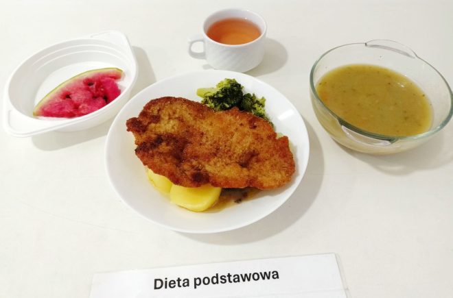 Na zdjęciu znajduje się posiłek oznaczony jako "Dieta podstawowa". Na talerzu umieszczono kotlet schabowy panierowany, podany z gotowanymi ziemniakami oraz brokułami. Obok talerza stoi miska z zupą, prawdopodobnie z ziemniaków lub warzyw. Po lewej stronie znajduje się miseczka z kawałkiem arbuza, a obok niej kubek z różowym napojem, przypuszczalnie kompotem