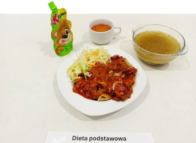 Na zdjęciu przedstawiono posiłek opisany jako "dieta podstawowa". Składa się z kilku elementów. Na talerzu znajduje się danie główne składające się z mięsa w sosie pomidorowym oraz surówki z kapusty. Obok talerza znajduje się miseczka z bulionem lub rosołem, filiżanka z herbatą oraz saszetka z napojem dla dzieci w kształcie misia. Na stole widnieje etykieta z napisem "Dieta podstawowa"