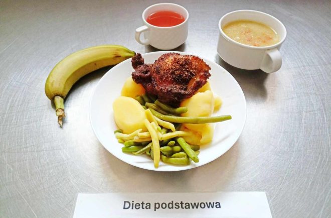 Na zdjęciu przedstawiono posiłek oznaczony jako "Dieta podstawowa". Na talerzu znajduje się porcja gotowanych ziemniaków, gotowane fasolki szparagowej (zielonej i żółtej) oraz smażony kawałek mięsa, przypominający kotlet. Obok na tacy znajduje się banan, filiżanka z czerwonym napojem, prawdopodobnie kompotem, oraz miseczka z zupą jarzynową