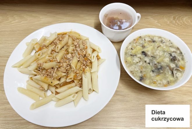 Na drugim zdjęciu widoczny jest obiad oznaczony jako "Dieta cukrzycowa". Na talerzu umieszczono makaron penne z sosem mięsnym, obok którego stoi kubek z herbatą oraz miska z gęstą zupą warzywną, prawdopodobnie z dodatkiem grzybów i ryżu