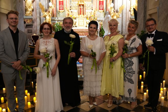 Fotografia przedstawia grupę elegancko ubranych osób - kobiet i mężczyzn, pozujących do zdjęcia w kościele na tle ołtarza. Wszystkie osoby trzymają w rękach po jednej, białej róży. Wśród osób znajduje się brat zakonny. Przed ołtarzem widać rozstawione, zapalone świece.