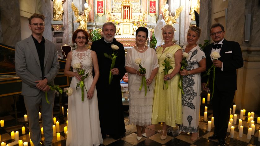 Fotografia przedstawia grupę elegancko ubranych osób - kobiet i mężczyzn, pozujących do zdjęcia w kościele na tle ołtarza. Wszystkie osoby trzymają w rękach po jednej, białej róży. Wśród osób znajduje się brat zakonny. Przed ołtarzem widać rozstawione, zapalone świece.