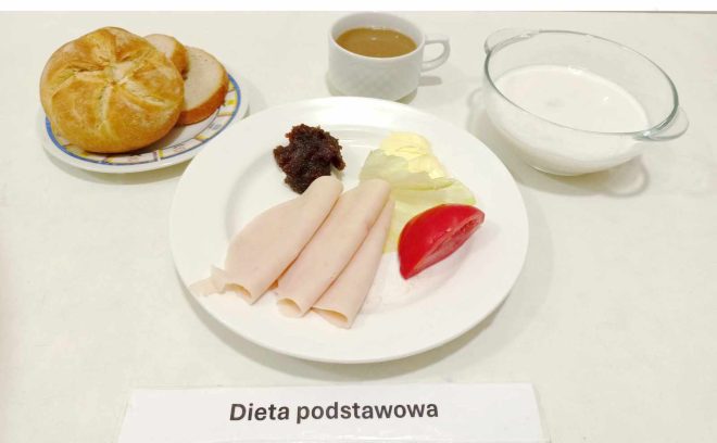 Taca z posiłkiem oznaczona jako "Dieta podstawowa". Na białym talerzu ułożono plastry wędliny, kawałek sałaty, plaster pomidora oraz porcję pasty lub dżemu. Obok na talerzu znajdują się bułka oraz dwa plastry chleba. Na stole znajduje się również filiżanka z kawą oraz miska z białym napojem, prawdopodobnie mlekiem.