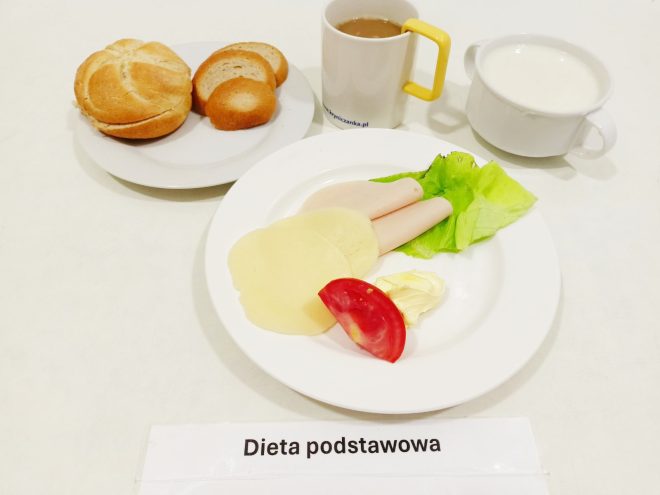 Na zdjęciu przedstawiono śniadanie oznaczone jako "Dieta podstawowa". Na talerzu znajdują się dwa plasterki wędliny drobiowej, dwa plasterki sera żółtego, plasterek pomidora oraz porcja masła i liść sałaty. Obok talerza umieszczono bułkę, dwie kromki chleba oraz dwa kubki: jeden z napojem (kawą z mlekiem lub herbatą), a drugi z białym płynem, prawdopodobnie kefirem, jogurtem lub mlekiem