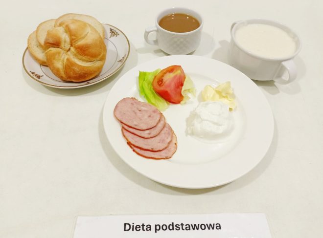 Na zdjęciu przedstawiono posiłek opisany jako "dieta podstawowa". Składa się z kilku elementów ułożonych na stole. Na talerzu znajduje się bułka kajzerka oraz dwa plasterki chleba. Na drugim talerzu znajduje się kilka plasterków wędliny, kawałek pomidora na liściu sałaty, porcja twarogu, trochę masła oraz kawałek czegoś białego (może to być jogurt lub inny nabiał). Obok talerzy znajduje się filiżanka z kawą oraz kubek z mlekiem lub białą kawą. Na stole widnieje etykieta z napisem "Dieta podstawowa"