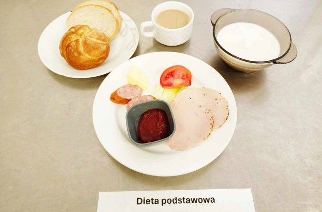 Na zdjęciu widoczny jest posiłek oznaczony jako "Dieta podstawowa". Na talerzu znajdują się plasterki wędliny i kiełbasy, porcja masła, plasterek pomidora, liść sałaty oraz mały pojemnik z dżemem. Obok na osobnym talerzyku leżą dwie kromki chleba i jedna bułka. Posiłek uzupełnia filiżanka z kawą z mlekiem oraz miseczka z białym napojem, prawdopodobnie mlekiem