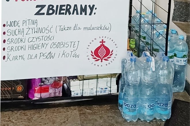 Zdjęcie przedstawia punkt zbiórki darów dla powodzian. Na plakacie znajdującym się w widocznym miejscu widnieje napis „Pomoc dla powodzian” oraz lista rzeczy, które można przynieść: woda pitna, sucha żywność (także dla małych dzieci), środki czystości, środki higieny osobistej oraz karma dla psów i kotów. Obok plakatu widać zgrzewki butelkowanej wody oraz różne inne produkty, w tym karmę dla zwierząt, ustawione na podłodze w metalowej kratce. Na plakacie znajduje się również logo Zakonu Szpitalnego św. Jana Bożego