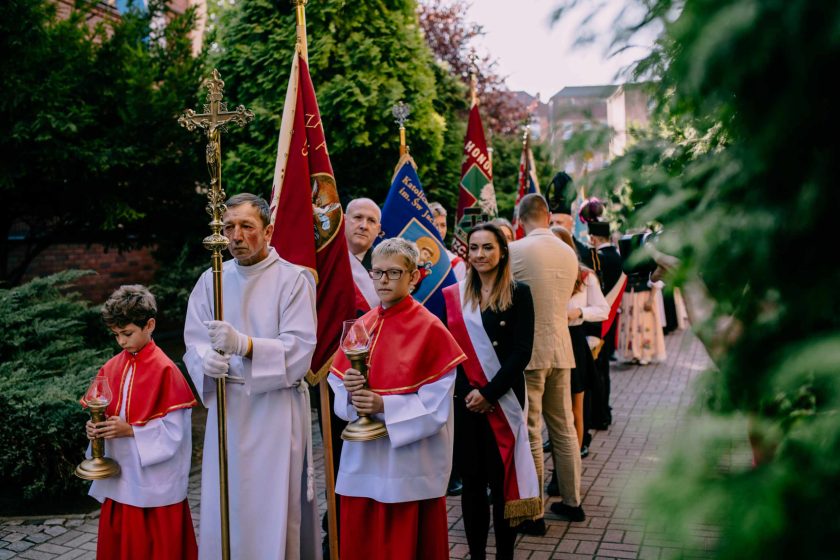 Procesja religijna na zewnątrz kościoła. Ministranci w czerwonych szatach liturgicznych niosą krzyż i świeczniki, a za nimi idzie grupa wiernych z różnymi sztandarami religijnymi, ubranymi w odświętne stroje