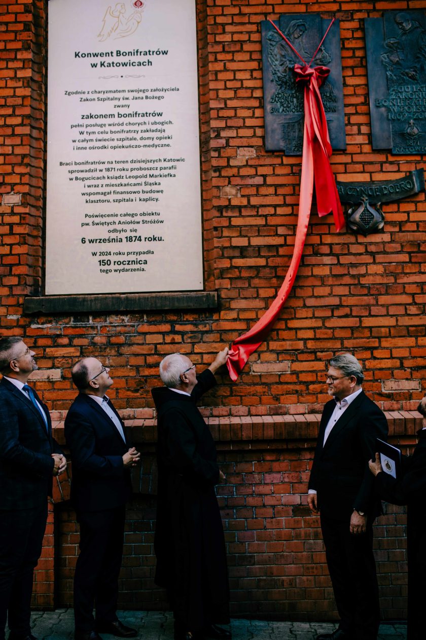 Uroczyste odsłonięcie tablicy upamiętniającej Bonifratrów na murze z cegły. Zakonnicy wraz z przedstawicielami duchowieństwa oraz świeckimi obserwują moment symbolicznego przecięcia wstęgi