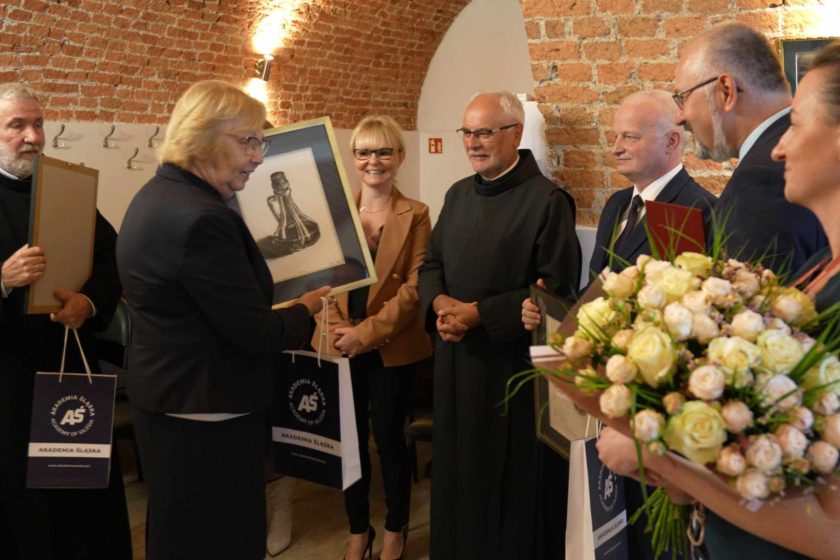 Starsza kobieta odbiera od zakonnika w czarnym habicie oprawiony obraz, uśmiechając się. W tle widać kilka osób, które również biorą udział w uroczystości