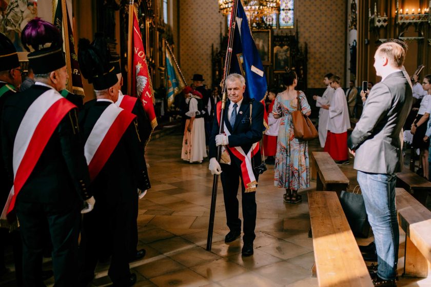 Scena wewnątrz kościoła, gdzie starsi mężczyźni w galowych uniformach i z szarfami stoją w rzędzie, trzymając sztandary, przygotowując się do ceremonii. W tle widać innych wiernych, z których część stoi, a część siedzi