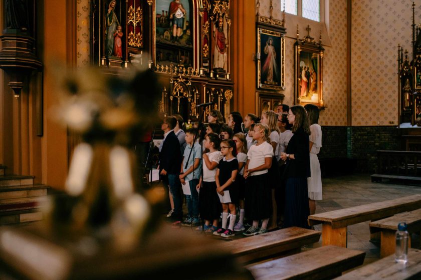 Dziecięcy chór ubrany w białe i ciemne stroje stoi przy ołtarzu w kościele, śpiewając podczas mszy lub innego wydarzenia religijnego. W tle widoczne są ozdobne obrazy sakralne na ścianach