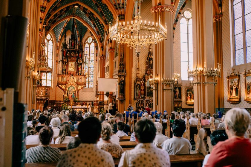 Wnętrze bogato zdobionego kościoła z tłumem wiernych siedzących w ławkach. W tle widoczny ołtarz z pięknymi witrażami, złoceniami oraz ozdobnymi obrazami religijnymi. Żyrandol wiszący pod gotyckimi łukami dodaje elegancji scenerii