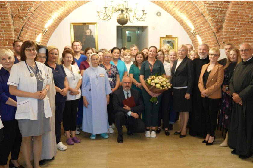 Grupa pracowników medycznych oraz zakonników pozujących razem w historycznym wnętrzu z ceglanymi sklepieniami. Na zdjęciu widoczne są osoby ubrane w medyczne uniformy oraz habit zakonny, a także osoby trzymające kwiaty