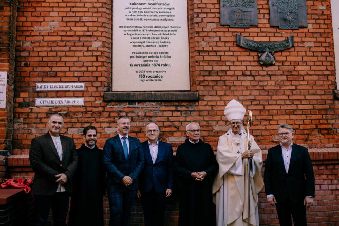 Grupa osób duchownych i świeckich pozujących do pamiątkowego zdjęcia przy tablicy na ceglanym murze. Na tablicy widnieje informacja o 150-leciu posługi Bonifratrów w Katowicach