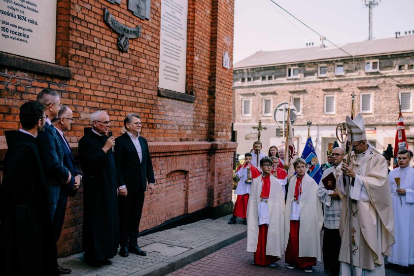 Duchowni i świeccy zebrani przy murze podczas uroczystości z udziałem procesji ministrantów. W tle widać tablice pamiątkowe i napis informujący o jubileuszu Bonifratrów