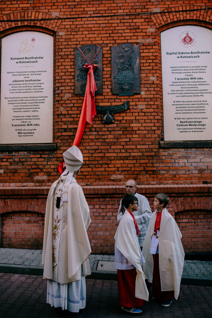 Na zdjęciu widzimy biskupa ubrany w liturgiczne szaty, stojącego przed ceglaną ścianą budynku, na której znajdują się tablice upamiętniające działalność Zakonu Bonifratrów w Katowicach. W pobliżu stoją ministranci w białych komżach i czerwonych albach, a całość otacza atmosfera uroczystości religijnej, związanej z jubileuszem 150-lecia Szpitala Zakonu Bonifratrów