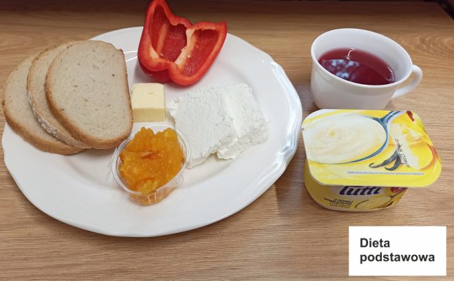 Na zdjęciu widoczny jest zestaw śniadaniowy oznaczony jako "Dieta podstawowa". Na talerzu znajdują się trzy kromki białego chleba, plaster masła, serek twarogowy, połowa czerwonej papryki oraz mała porcja dżemu. Obok talerza znajduje się kubek z herbatą owocową i jogurt o smaku waniliowym