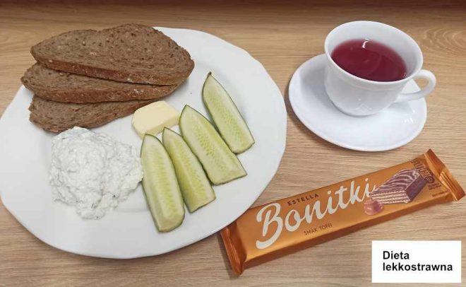 Na talerzu widzimy trzy kromki ciemnego chleba, porcję twarogu, plasterek masła oraz kilka kawałków świeżego ogórka. Obok talerza leży batonik "Bonitki" o smaku toffi oraz filiżanka z różowym napojem, prawdopodobnie herbatą. Taca ma etykietę z napisem "Dieta lekkostrawna"