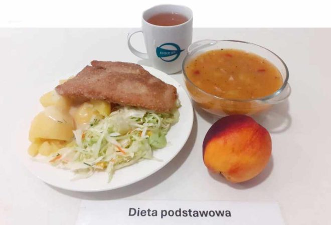 Na zdjęciu znajduje się posiłek oznaczony jako "Dieta podstawowa". Na talerzu umieszczono panierowany kotlet, ziemniaki polane sosem oraz porcję surówki z białej kapusty. Obok widnieje przezroczysta miseczka z zupą jarzynową, filiżanka z herbatą oraz brzoskwinia