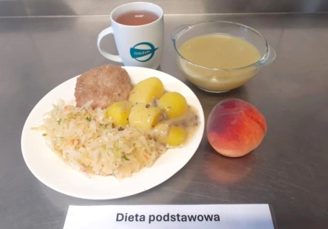 Na zdjęciu przedstawiono zestaw obiadowy oznaczony jako "Dieta podstawowa". Na talerzu znajduje się kotlet mielony, ziemniaki w sosie, a także porcja kiszonej kapusty. Obok znajduje się brzoskwinia, kubek z herbatą oraz miseczka z zupą