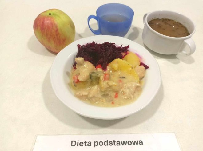 Na zdjęciu znajduje się posiłek oznaczony jako "Dieta podstawowa". W centralnej części widzimy głęboką miskę z daniem składającym się z duszonych warzyw i kawałków mięsa w sosie, obok porcji gotowanych ziemniaków i surówki z buraków. W tle po lewej stronie leży jabłko, a z prawej znajdują się dwa kubki – jeden z przezroczystą herbatą w niebieskim kubku, a drugi z ciemnym płynem przypominającym zupę lub bulion w białej filiżance