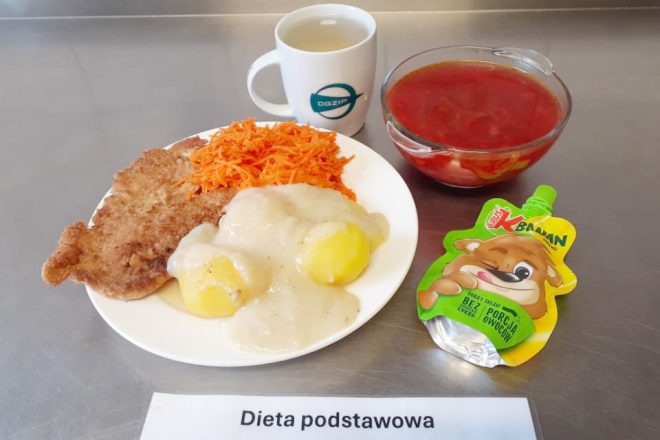 Zdjęcie przedstawia podstawowy obiad w szpitalu. Na talerzu znajdują się panierowany kotlet, ziemniaki polane sosem, oraz porcja tartej marchewki. Obok znajduje się miska z czerwonym barszczem oraz torebka musu owocowego o smaku banana. Podano także filiżankę herbaty. Na karteczce widnieje napis „Dieta podstawowa”