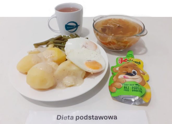 Na zdjęciu widoczny jest obiad oznaczony jako "Dieta podstawowa." Na białym talerzu znajdują się ziemniaki polane sosem, smażone jajko sadzone oraz porcja fasolki szparagowej. W tle znajduje się kubek z herbatą, miseczka z zupą oraz owocowy mus w tubce