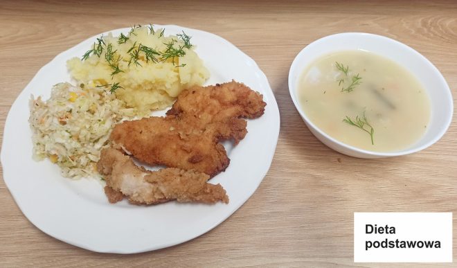 Zdjęcie przedstawia obiad na diecie podstawowej. Na talerzu znajdują się panierowane kawałki mięsa (kotlet), porcja puree ziemniaczanego posypana koperkiem, surówka z kapusty oraz miska zupy. Na kartce widnieje napis „Dieta podstawowa”